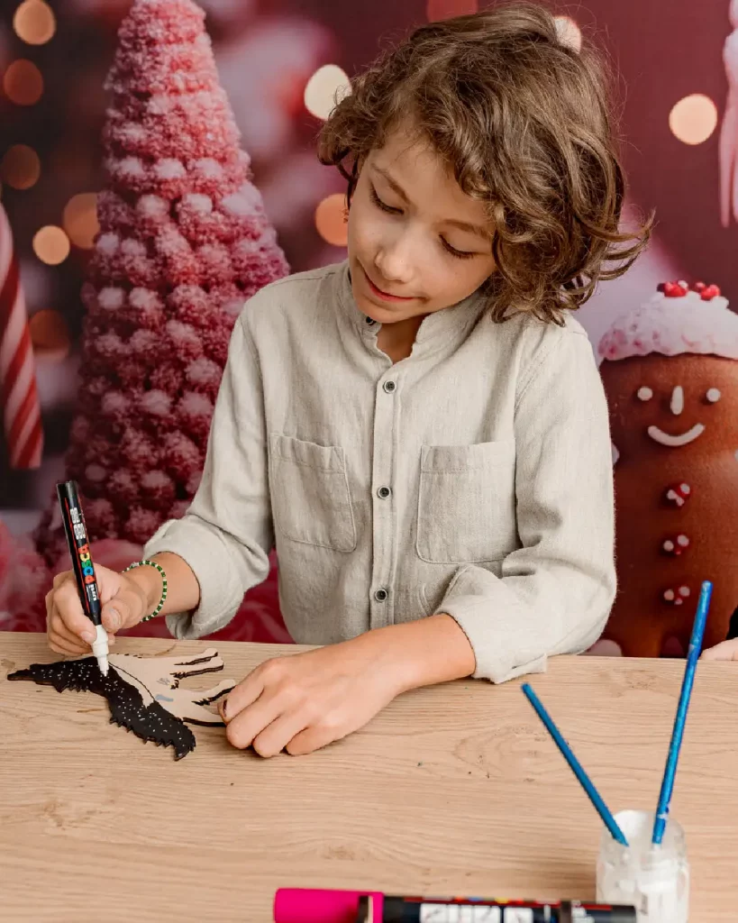 Un petit garçon qui décorent son loup en bois. Le loup contient un paysage de montagne découpés à l'intérieur (arbres, montagne). Il colle ensuite des mini-animaux en bois pour rendre la création unique. Ateliers de création en bois pour les familles dans les stations de ski et les offices de tourisme. Les enfants et les familles fabriquent un loup en bois composée de 4 pièces en bois et d'un socle. Cette animation convient aux offices de tourisme est accessibles aux enfants dès 6 ans et leurs familles ! Cet événement proposé par Les graines d'Eveil est accessible avec et sans inscription aux enfants à partir de 6 ans et leurs familles. Ils décorent et assemblent leurs création. Ils repartent avec un loup gravé avec le nom de l'événement ou de la station de ski. Cette animation est à destination des stations de Savoie et offices de tourisme (Courchevel, Tignes, Val d'Isère, Sainte Foy, La Rosière, Les Arcs, Peisey-Vallandry, La Plagne, Val Thorens) et Haute Savoie (Megève, Combloux, Chamonix, Morzine les Gets, Annecy, Avoriaz, La Cluzaz...). Il le personnalise avec des animaux en bois. C'est un souvenir unique et inoubliable de leurs vacances en stations de ski.