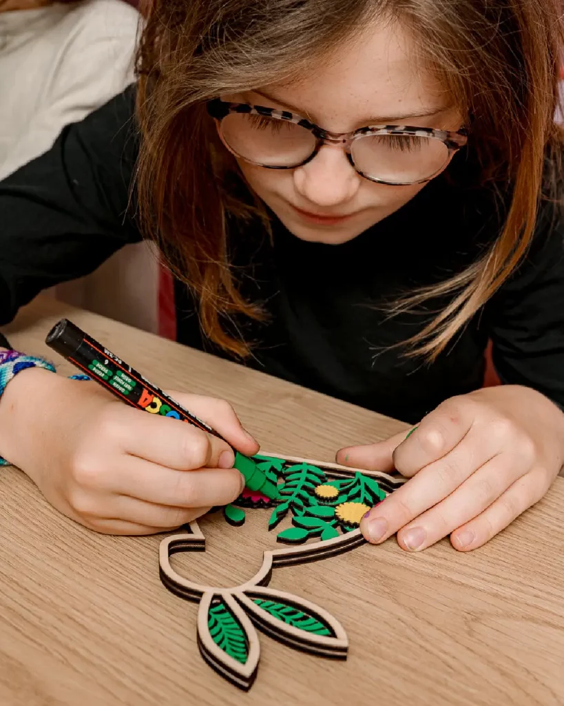 Une petite fille qui colorie les différentes couches de son lapin en bois. Le lapin contient un paysage de montagne découpés à l'intérieur (arbres, montagne). Elle colle ensuite des mini-animaux en bois pour rendre la création unique. Ateliers de création en bois pour les familles dans les stations de ski et les offices de tourisme. Les enfants et les familles fabriquent un lapin en bois composée de 4 pièces en bois et d'un socle. Cette animation convient aux offices de tourisme est accessibles aux enfants dès 6 ans et leurs familles ! Cet événement proposé par Les graines d'Eveil est accessible avec et sans inscription aux enfants à partir de 6 ans et leurs familles. Ils décorent et assemblent leurs création. Ils repartent avec un lapin gravé avec le nom de l'événement ou de la station de ski. Cette animation est à destination des stations de Savoie et offices de tourisme (Courchevel, Tignes, Val d'Isère, Sainte Foy, La Rosière, Les Arcs, Peisey-Vallandry, La Plagne, Val Thorens) et Haute Savoie (Megève, Combloux, Chamonix, Morzine les Gets, Annecy, Avoriaz, La Cluzaz...). Il le personnalise avec des animaux en bois. C'est un souvenir unique et inoubliable de leurs vacances en stations de ski.