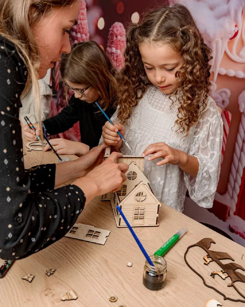 La petit fille colle les pièces de son chalet en bois et l'animatrice l'aide à construire sa création. Ateliers de création en bois pour les familles dans les stations de ski. Les enfants et les familles fabriquent un chalet en bois composée de 10 pièces en bois. Cette animation convient aux offices de tourisme est accessibles aux enfants dès 6 ans et leurs familles ! Cet événement est accessible avec et sans inscription aux enfants à partir de 6 ans et leurs familles. Ils décorent et assemblent leurs création. Ils repartent avec un chalet en bois gravés avec le nom de l'événement ou de la station de ski. Cette animation est à destination des stations de Savoie et offices de tourisme (Courchevel, Tignes, Val d'Isère, Sainte Foy, La Rosière, Les Arcs, Peisey-Vallandry, La Plagne, Val Thorens) et Haute Savoie (Megève, Combloux, Chamonix, Morzine les Gets, Annecy, Avoriaz, La Cluzaz...). Il le personnalise avec des mini-ski, des flocons, des étoiles, des sapins...