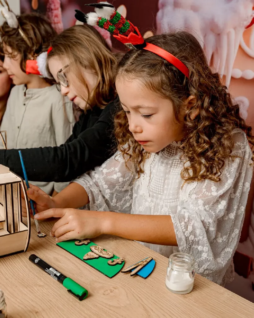 La petite fille décore et colle les mini-déorations (flocons, skis, étoiles..) de sa télécabine en bois avec de la peinture. Ateliers de création en bois pour les familles dans les stations de ski et les offices de tourisme. Les enfants et les familles fabriquent une télécabine en bois composée de 20 pièces en bois. Cette animation convient aux offices de tourisme est accessibles aux enfants dès 6 ans et leurs familles ! Cet événement proposé par Les graines d'Eveil est accessible avec et sans inscription aux enfants à partir de 6 ans et leurs familles. Ils décorent et assemblent leurs création. Ils repartent avec une télécabine gravés avec le nom de l'événement ou de la station de ski. Cette animation est à destination des stations de Savoie et offices de tourisme (Courchevel, Tignes, Val d'Isère, Sainte Foy, La Rosière, Les Arcs, Peisey-Vallandry, La Plagne, Val Thorens) et Haute Savoie (Megève, Combloux, Chamonix, Morzine les Gets, Annecy, Avoriaz, La Cluzaz...). Il le personnalise avec des mini-ski, des flocons, des étoiles, des sapins... Il utilise un fil en bois pour la suspendre à la maison. C'est un souvenir unique et inoubliable de leurs vacances en stations de ski.