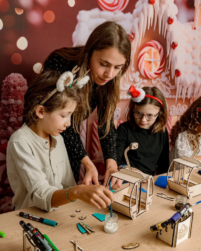 Le petit garçon colle les pièces de sa télécabine en bois et l'animatrice l'aide à assembler sa télécabine en bois. Ateliers de création en bois pour les familles dans les stations de ski et les offices de tourisme. Les enfants et les familles fabriquent une télécabine en bois composée de 20 pièces en bois. Cette animation convient aux offices de tourisme est accessibles aux enfants dès 6 ans et leurs familles ! Cet événement proposé par Les graines d'Eveil est accessible avec et sans inscription aux enfants à partir de 6 ans et leurs familles. Ils décorent et assemblent leurs création. Ils repartent avec une télécabine gravés avec le nom de l'événement ou de la station de ski. Cette animation est à destination des stations de Savoie et offices de tourisme (Courchevel, Tignes, Val d'Isère, Sainte Foy, La Rosière, Les Arcs, Peisey-Vallandry, La Plagne, Val Thorens) et Haute Savoie (Megève, Combloux, Chamonix, Morzine les Gets, Annecy, Avoriaz, La Cluzaz...). Il le personnalise avec des mini-ski, des flocons, des étoiles, des sapins... Il utilise un fil en bois pour la suspendre à la maison. C'est un souvenir unique et inoubliable de leurs vacances en stations de ski