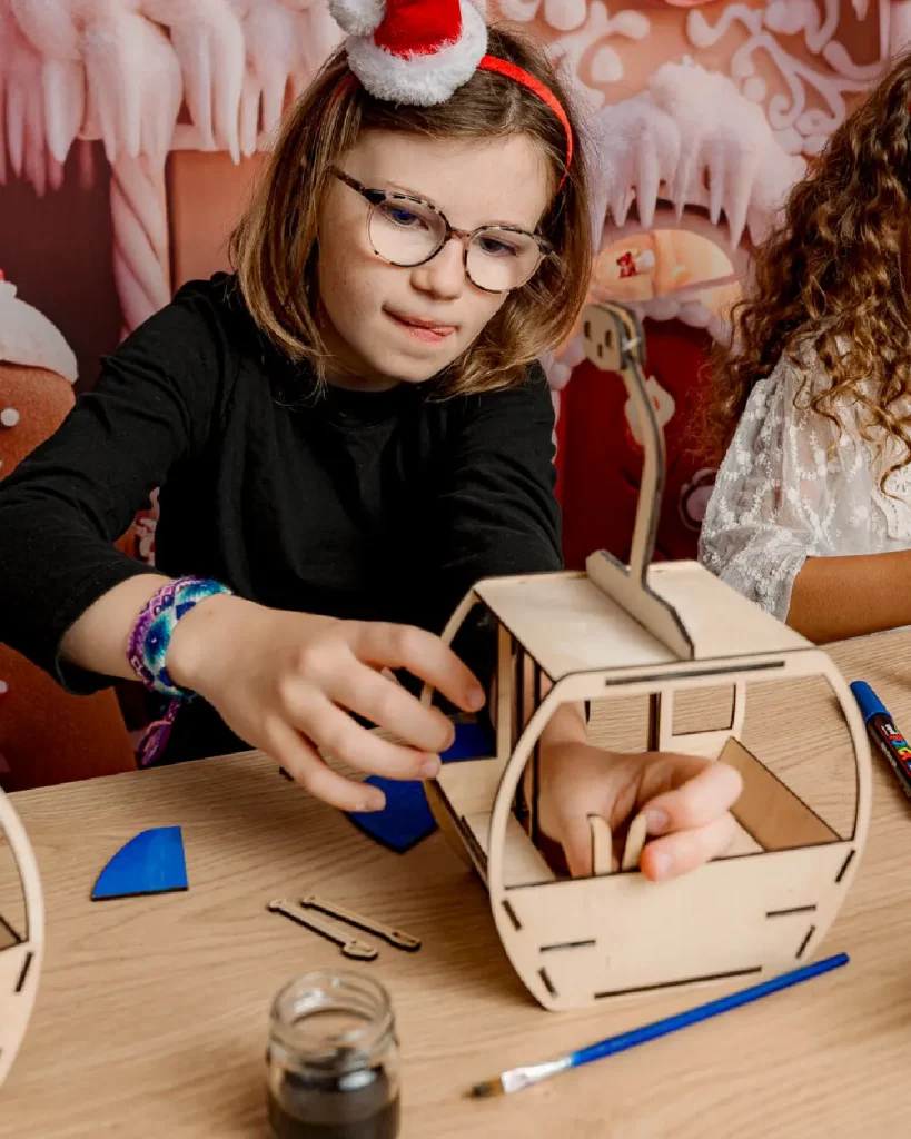 La petit fille colle les pièces de sa télécabine en bois. Ateliers de création en bois pour les familles dans les stations de ski et les offices de tourisme. Les enfants et les familles fabriquent une télécabine en bois composée de 20 pièces en bois. Cette animation convient aux offices de tourisme est accessibles aux enfants dès 6 ans et leurs familles ! Cet événement est accessible avec et sans inscription aux enfants à partir de 6 ans et leurs familles. Ils décorent et assemblent leurs création. Ils repartent avec une télécabine gravés avec le nom de l'événement ou de la station de ski. Cette animation est à destination des stations de Savoie et offices de tourisme (Courchevel, Tignes, Val d'Isère, Sainte Foy, La Rosière, Les Arcs, Peisey-Vallandry, La Plagne, Val Thorens) et Haute Savoie (Megève, Combloux, Chamonix, Morzine les Gets, Annecy, Avoriaz, La Cluzaz...). Il le personnalise avec des mini-ski, des flocons, des étoiles, des sapins... Il utilise un fil en bois pour la suspendre à la maison. C'est un souvenir unique et inoubliable de leurs vacances en stations de ski
